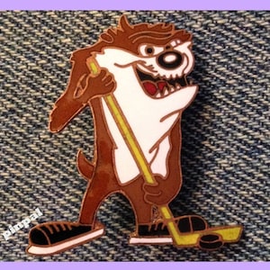 Pode incluir: Um Diabo da Tasmânia de desenho animado marrom e branco vestindo um patim de hóquei preto e segurando um taco de hóquei amarelo. O personagem está em pé em um fundo de jeans azul.