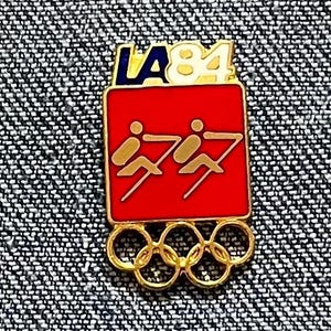May include: A vintage pin featuring the LA 84 logo in blue and white. The pin has a red square with gold figures and the Olympic rings in gold at the bottom. The pin is on a textured background.