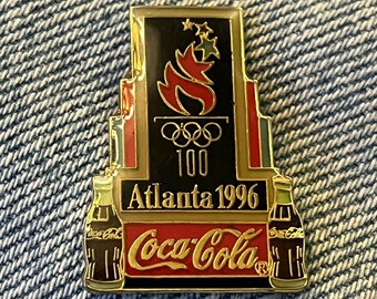 May include: A rectangular Coca-Cola pin featuring the Atlanta 1996 Olympics logo. The pin has a black background with a red and gold border, Olympic rings, and two Coca-Cola bottle designs.