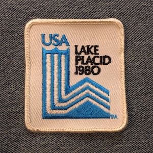 May include: A white embroidered patch with the text "USA" and a blue and white stylized building. The text "LAKE PLACID 1980" is also on the patch.