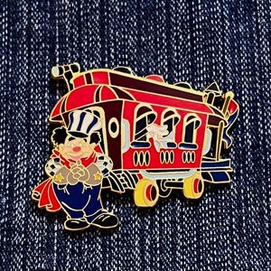 Könnte beinhalten: Ein farbenfroher Anstecker mit einer Zeichentrickfigur in einer Schaffneruniform neben einem roten Waggon. Der Waggon hat goldene Akzente und die Nummer "000" auf der Seite. Der Anstecker ist vor einem dunkelblauen Jeanshintergrund platziert.