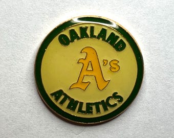 May include: A circular pin featuring the Oakland Athletics logo. The pin has a yellow background with a large yellow "A" and the words "OAKLAND" and "ATHLETICS" in green. The pin is trimmed with a green border.
