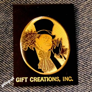 May include: A gold-colored pin featuring a cartoon character with a top hat, curly hair, and a monocle. The character is wearing a black suit and a white bow tie. The pin is on a black background with the text "GIFT CREATIONS, INC."
