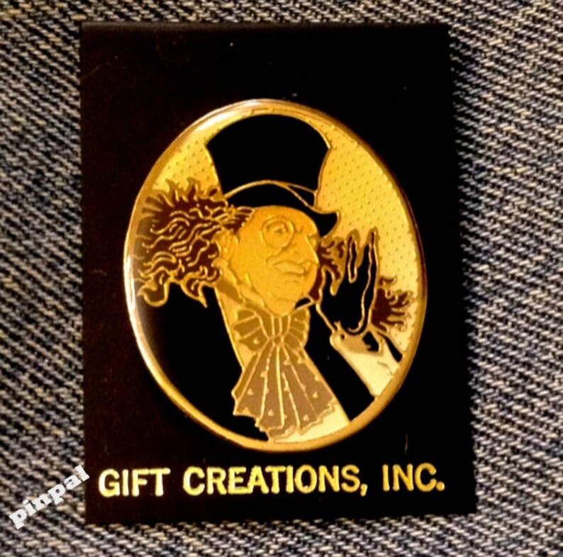 May include: A gold-colored pin featuring a cartoon character with a top hat, curly hair, and a monocle. The character is wearing a black suit and a white bow tie. The pin is on a black background with the text "GIFT CREATIONS, INC."