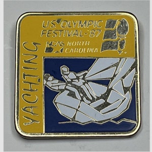 May include: A gold-colored pin with a blue and white image of a sailboat. The pin reads "US Olympic Festival '87" and "NCAS North Carolina" in gold lettering.
