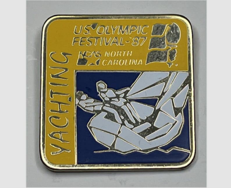 May include: A gold-colored pin with a blue and white image of a sailboat. The pin reads "US Olympic Festival '87" and "NCAS North Carolina" in gold lettering.