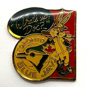 May include: A vintage pin featuring Bugs Bunny in a baseball uniform holding a bat, with the text "What's up, Doc?" and the Toronto Blue Jays logo. The pin has a gold border and a red background.