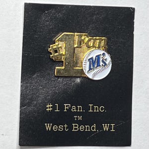 May include: A gold-colored pin with the words "#1 Fan" and a baseball design. The pin features the letter "M" in blue on a white baseball. The pin is on a black card with text that reads "#1 Fan, Inc." and "West Bend, WI."