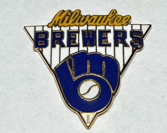 May include: A Milwaukee Brewers baseball team pin. The pin features the team's logo, including the words "Milwaukee Brewers" in yellow and blue, and a blue baseball glove with a white baseball.