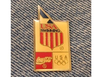 May include: A rectangular gold-colored pin featuring the text "USA Swimming" above a red and white striped shield. The pin also includes the Coca-Cola logo and the Olympic rings, with the text "USA".