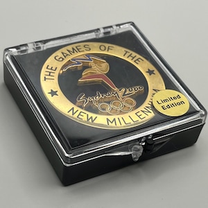 Peut inclure: Une pièce de monnaie commémorative dorée des Jeux olympiques de Sydney 2000, présentée dans un étui en plastique transparent avec une base noire. La pièce porte les anneaux olympiques et le texte "The Games of the New Millennium". Un petit autocollant indique "Limited Edition".