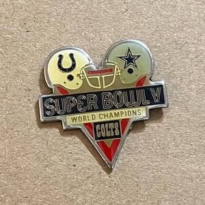Puede incluir: Un pin en forma de corazón con dos cascos de fútbol americano, uno con una herradura y otro con una estrella. El pin dice "SUPER BOWL V WORLD CHAMPIONS COLTS" en letras negras y rojas, con un borde plateado.