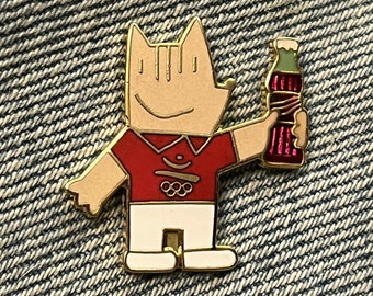 May include: Gold-colored pin featuring a cartoon character holding a red and white Coca-Cola bottle. The character is wearing a red shirt with the Olympic rings and white pants.