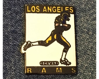 May include: A gold and black Los Angeles Rams pin featuring a cartoon football player running with the ball. The pin says "Los Angeles Rams" and "IGHSWAD" in gold lettering.