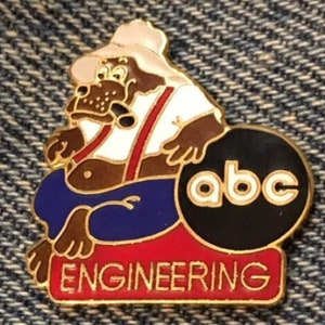 Op de afbeelding: Een gouden speld met een cartoonfiguur dat een wit shirt, een overall en een hoed draagt. Het figuur zit op een rood en blauw kussen naast een zwarte cirkel met de letters "abc" in wit. De tekst "ENGINEERING" staat in gouden letters op een rode achtergrond onder het figuur.