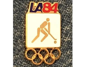 May include: Gold-colored pin with a white background featuring a stick figure playing field hockey. The pin also features the text "LA84" in blue and red letters and five interlocking gold rings.