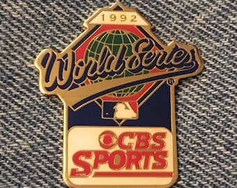 May include: A gold-colored pin with a blue and white baseball diamond design. The pin features the text "World Series" in a gold script font, a globe, and the text "CBS SPORTS" in a red and white font. The year "1992" is printed in red at the top of the pin.