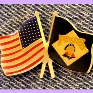 Puede incluir: Un pin de color dorado con dos banderas cruzadas. La bandera de la izquierda es la bandera estadounidense con rayas rojas y blancas y estrellas azules. La bandera de la derecha es negra con un rombo blanco con la palabra "Pirates" en letras doradas. El rombo también tiene un pirata de dibujos animados con un sombrero dorado y un pañuelo rojo.
