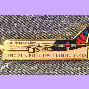 May include: A gold-colored pin featuring a Delta Air Lines airplane with the text "Delta Air Lines" and "Spirit of Delta". The airplane is flying over a blue and red background with the text "Atlanta 1996" and the Olympic rings. The pin also has the text "Official Airline 1996 Olympic Games".