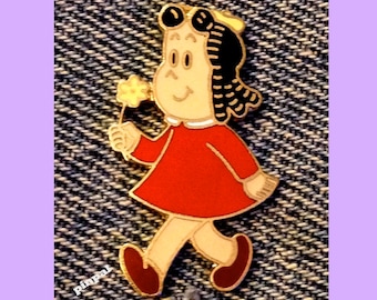May include: A gold-colored pin featuring a cartoon girl with black hair, wearing a red dress and holding a yellow flower.