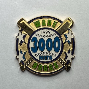 Peut inclure: Épinglette dorée sur le thème du baseball. L'épinglette présente le texte "WADE BOGGS" et "3000 HITS 1999" en vert et bleu. Deux battes de baseball croisées et quatre étoiles blanches avec des accents bleus sont également présentes.