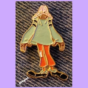 May include: A gold-coloured enamel pin featuring a cartoon character wearing a pink and white hat, a green sweater with black stripes, orange trousers, and black shoes.