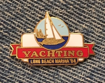 May include: Gold-colored pin with a red, white, and blue design. The pin features a sailboat on a blue and white background with the text "YACHTING LONG BEACH MARINA '84".