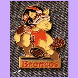 Peut inclure: Une épingle dorée représentant un cheval de dessin animé portant un casque de football américain et un maillot. Le cheval tient un ballon de football et l'épingle porte l'inscription "Broncos" en bas.