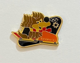 May include: A gold-toned pin featuring the Sarajevo '84 Olympic mascot, a cartoon wolf on a sled. The wolf has brown fur, a red scarf, and a yellow face. The pin includes the Olympic rings and the text "Sarajevo '84".