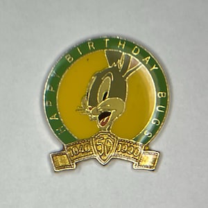 Puede incluir: Un pin de color dorado con un borde verde. El pin presenta una imagen de dibujos animados de Bugs Bunny con el texto "Happy Birthday Bugs 1940 50 1990" alrededor del borde.
