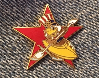 May include: A red star-shaped pin with a cartoon eagle wearing a red, white, and blue hat paddling a yellow kayak. The eagle is holding a white paddle.