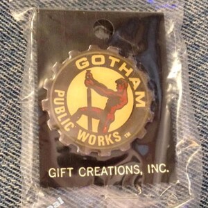 May include: A gold and black pin with a gear design. The pin features a cartoon image of a man working on a construction project. The text on the pin reads "Gotham Public Works". The pin is packaged in a clear plastic bag with a black card that reads "Gift Creations, Inc.".