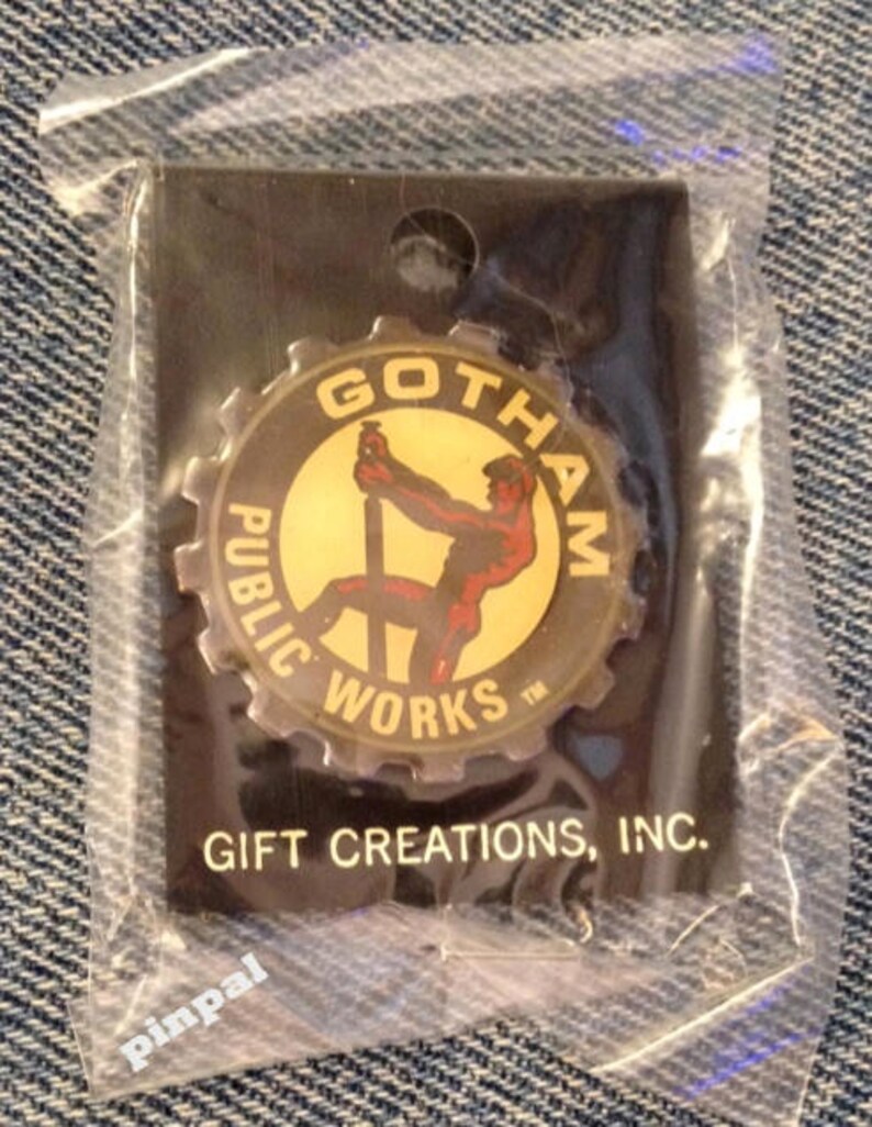 May include: A gold and black pin with a gear design. The pin features a cartoon image of a man working on a construction project. The text on the pin reads "Gotham Public Works". The pin is packaged in a clear plastic bag with a black card that reads "Gift Creations, Inc.".