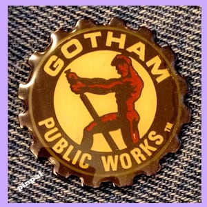 May include: A round, brown metal pin with a yellow and red image of a man working on a construction project. The pin says "Gotham Public Works" in a circle around the image.