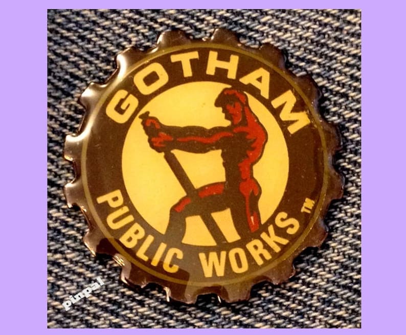 May include: A round, brown metal pin with a yellow and red image of a man working on a construction project. The pin says "Gotham Public Works" in a circle around the image.