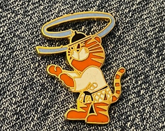 May include: A gold-colored pin featuring a cartoon tiger wearing a white karate uniform and a blue headband. The tiger is holding a blue and white ribbon in its paws.