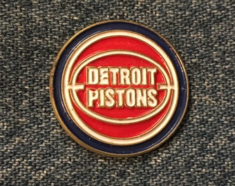 May include: A red, white, and blue enamel pin with the words "DETROIT PISTONS" in white lettering inside a basketball shape.