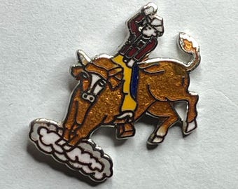 May include: A detailed enamel pin depicting a rodeo scene. A cowboy in a red jacket and white hat rides a bucking bull. The bull is brown with white accents, and the pin has a silver border and a cloud-like design at the bottom.
