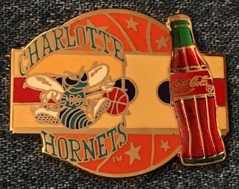 May include: A gold-colored pin featuring the Charlotte Hornets logo, a cartoon hornet dribbling a basketball, with the words "Charlotte Hornets" and a Coca-Cola bottle.