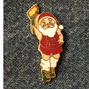 May include: A gold-coloured pin featuring a cartoon Santa Claus wearing a red and white athletic uniform with the text "L.A. '84". He is holding a torch in his right hand and has a white beard and moustache.