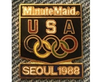 May include: A gold-colored pin with a black shield-shaped background. The shield has the text "Minute Maid" at the top, "USA" in the center, and five interlocking Olympic rings in gold, red, blue, green, and black. The bottom of the shield has a gold banner with the text "Seoul 1988".