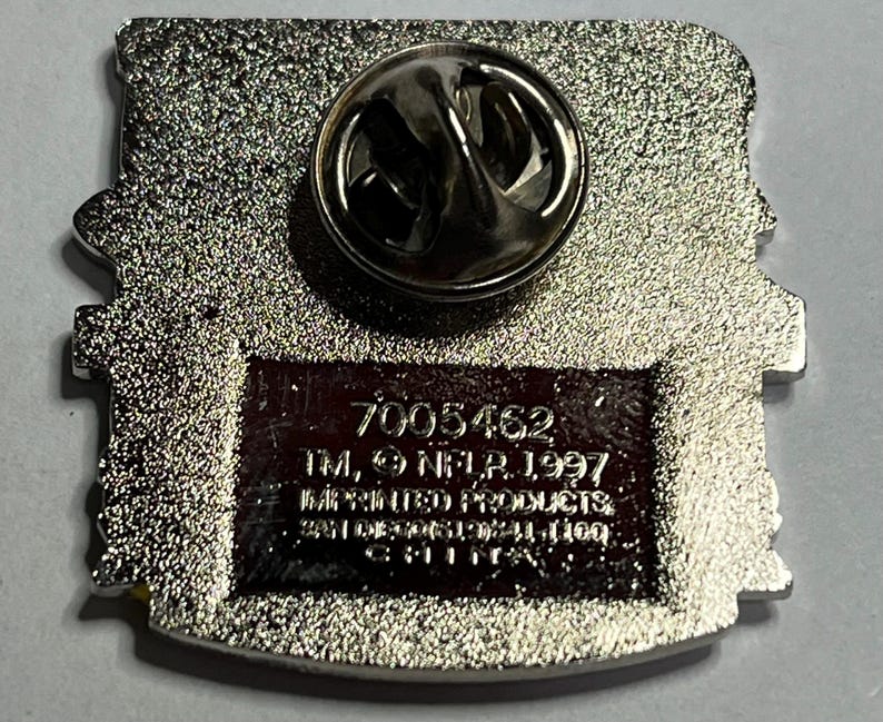 May include: A silver-colored pin with a pin back. The pin has a rectangular shape with rounded edges. The back of the pin has the text "7005462 TM, NFLP, 1997, IMPRINTED PRODUCTS, 2010 NFL PROPERTIES LLC, CHINA."