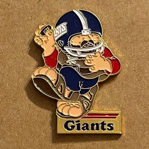 May include: A colourful enamel pin depicting a cartoon football player in a blue helmet with the word "Giants" on the helmet and the base of the pin. The player wears a red and brown uniform.