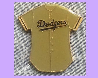 May include: A gold-colored baseball jersey pin with the word "Dodgers" in black lettering across the front.