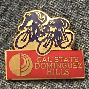 Könnte beinhalten: Eine goldene Anstecknadel mit einer blauen Silhouette von zwei Radfahrern im Rennen. Die Anstecknadel hat einen roten Hintergrund mit dem Text "CAL STATE DOMINGUEZ HILLS" in goldenen Buchstaben.