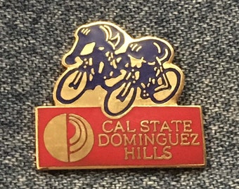 May include: A gold pin with a blue silhouette of two cyclists racing. The pin has a red background with the text "CAL STATE DOMINGUEZ HILLS" in gold lettering.