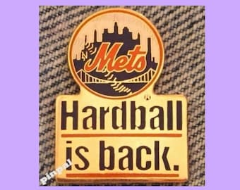 May include: A gold-colored pin with a blue and white New York Mets logo and the text "Hardball is back." The logo features a stylized image of the New York City skyline and the Brooklyn Bridge.