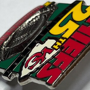 May include: A metal pin with a red, green, and yellow color scheme. The pin features the text "25TH CHIEFS" in red and yellow, along with a logo. The pin has a silver border and a red design on the left side.