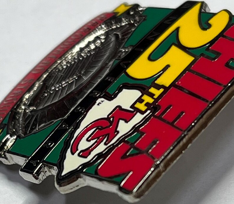 May include: A metal pin with a red, green, and yellow color scheme. The pin features the text "25TH CHIEFS" in red and yellow, along with a logo. The pin has a silver border and a red design on the left side.