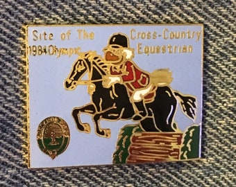 May include: A gold-colored pin with a blue background. The pin features a cartoon image of a rider on a horse jumping over a fence. The text "Site of The 1984 Olympic Cross-Country Equestrian" is printed on the pin.
